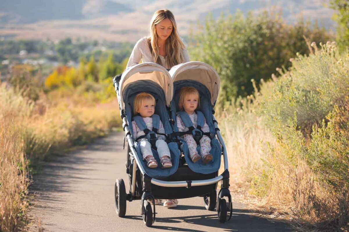 A woman with long blonde hair pushes two young children in a side-by-side double stroller.