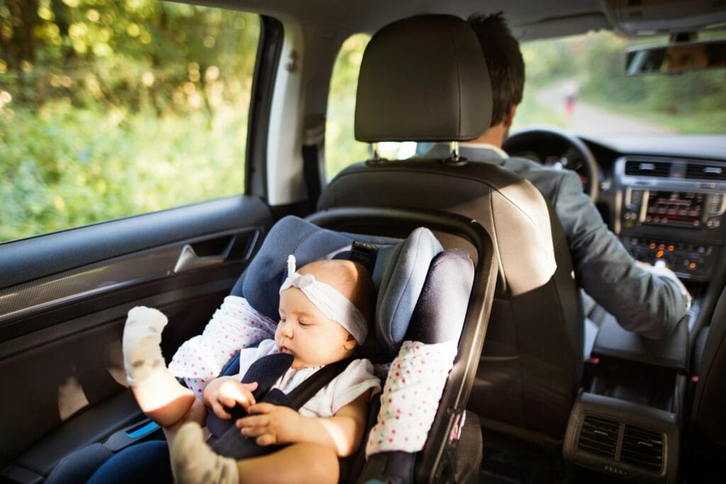 A baby is secured in an infant car seat in the back of a car while a man sits in the driver's seat.