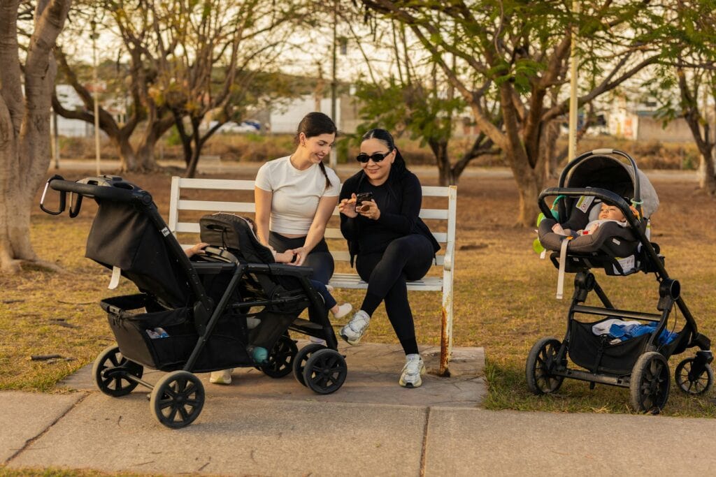 Two women are sitting on a park bench, looking at a smartphone. A black double stroller and a stroller with a car seat attached are parked beside them. The image illustrates the Benefits of Using a Double Stroller for convenient outdoor activities